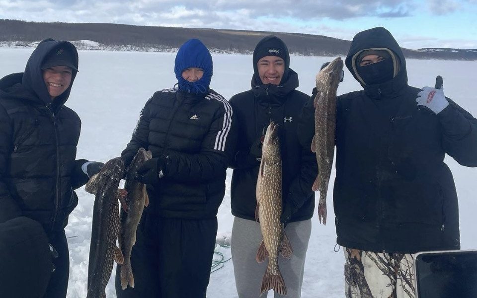 Four Indigenous people wearing winter clothing stand outside holding fish