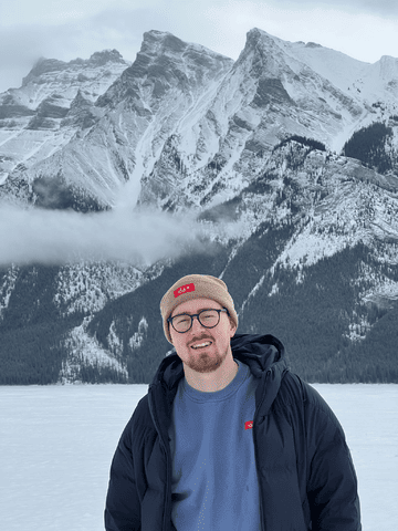 A man wearing a toque and glasses stands smiling in front of the mountains
