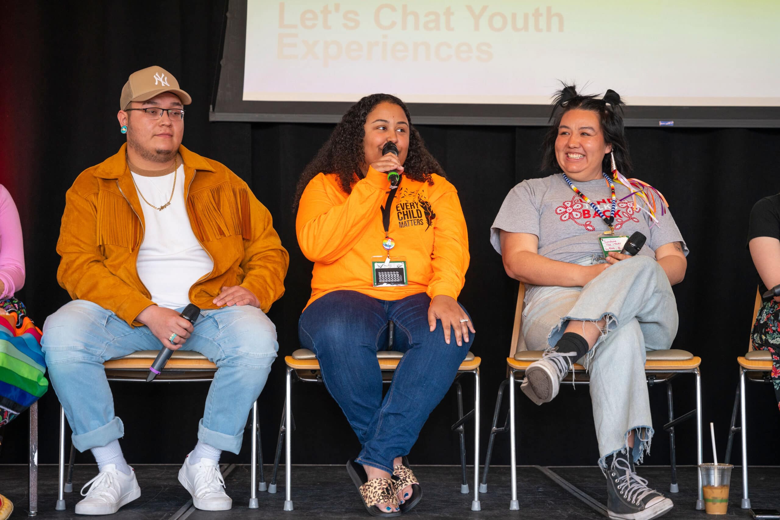 Three Indigenous youth sit on a stage giving a presentation. One is male and wears a hide jacket, one is Afro-Indigenous and wears an orange 'Every Child Matters' shirt, and one wears a grey shirt