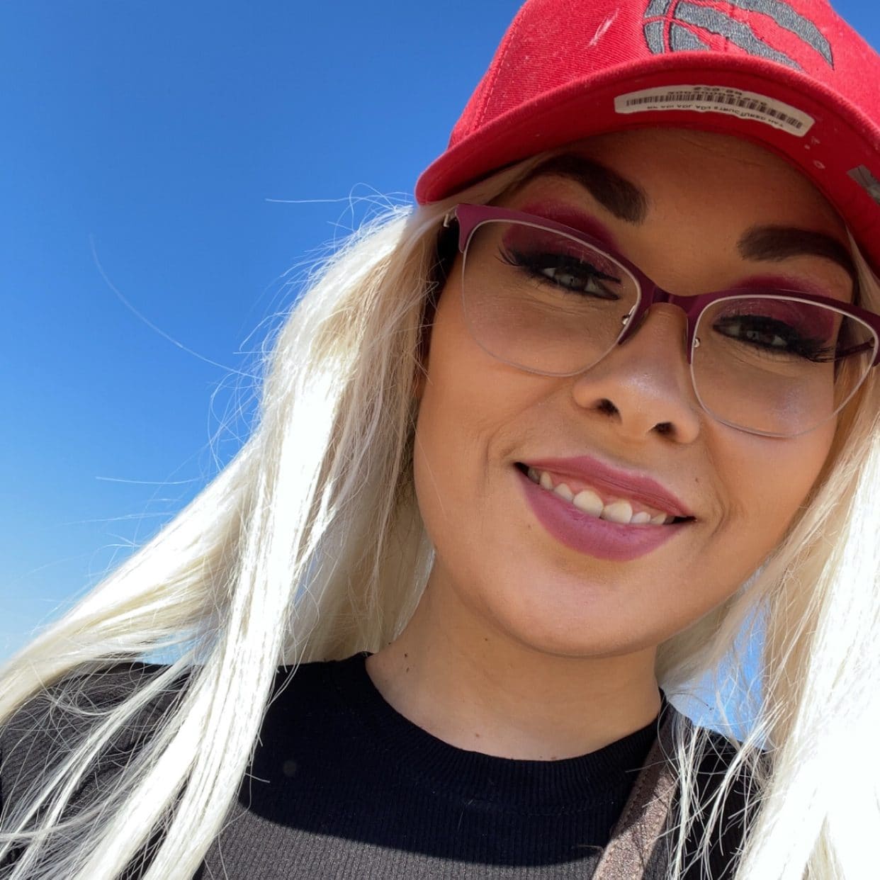 A young woman with long blonde hair wearing a red hat and glasses smiles at the camera