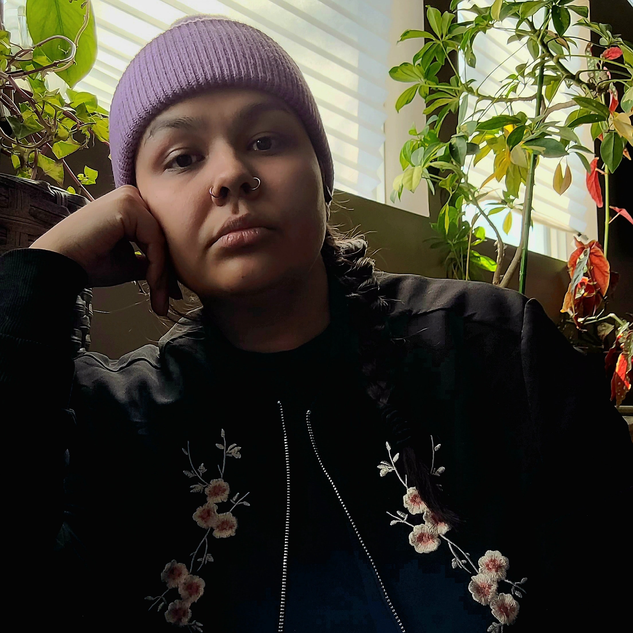 A person wearing a purple hat and a bomber jacket with flowers on it looks into a camera
