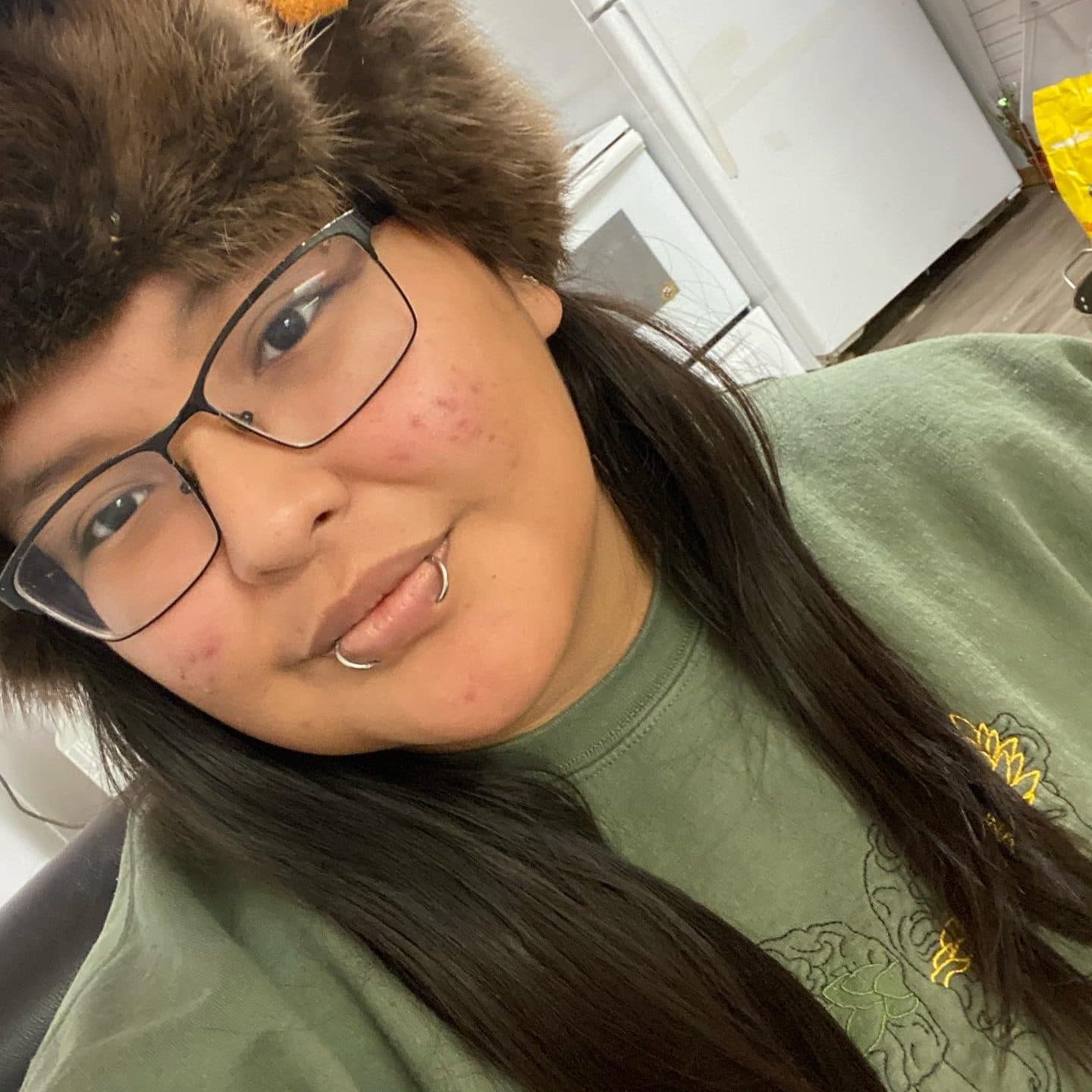 A person with long brown hair wearing a fur hat and glasses smils for the camera