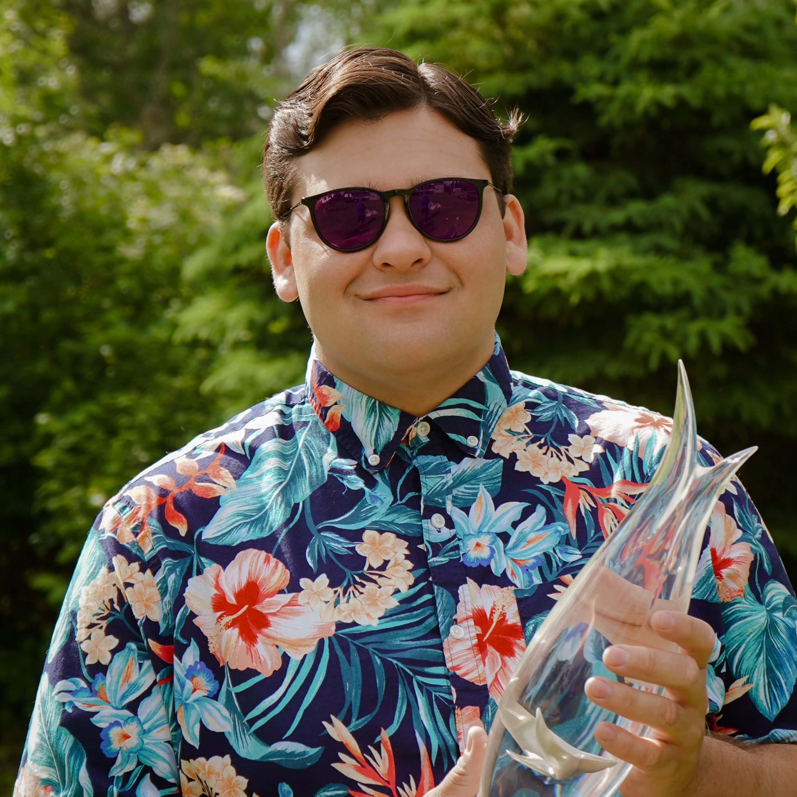 A young man with brown hair wearing a blue floral print shirt and sunglasses poses for a photo
