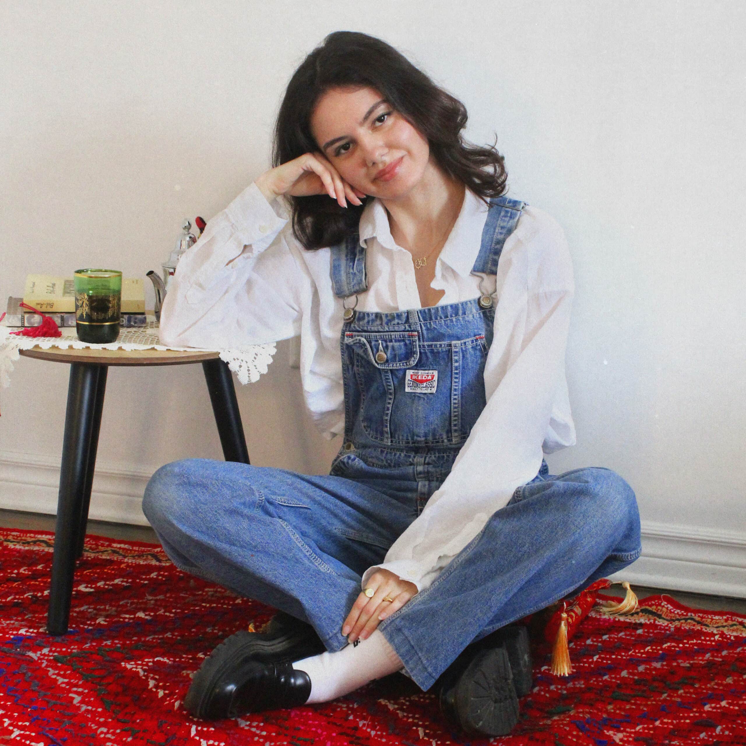 Amani smiles is sitting cross legged on a red rug smiling at the camera. She is wearing a white dress shirt with blue overalls.