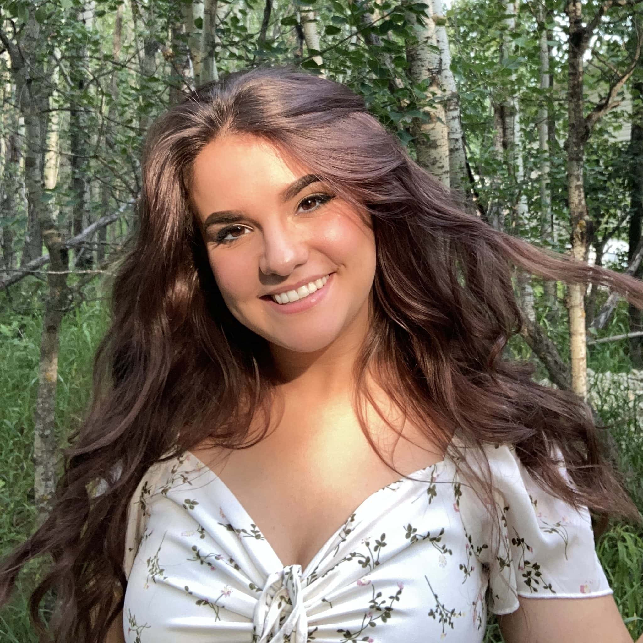 A young person with long brown hair smiling and standing in a forest