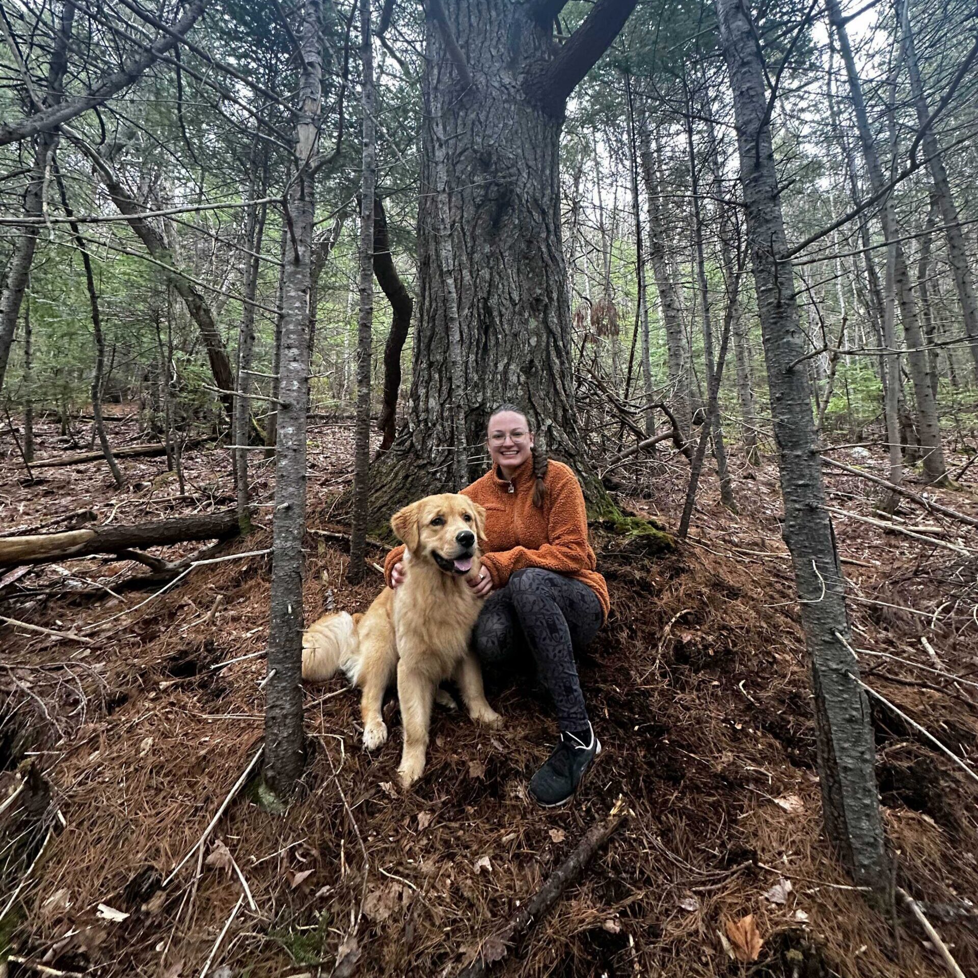 Sabrina is sitting next to her Golden Retriever puppy, at the base of an old Balsam Fir tree. They're in a densely wooded area.