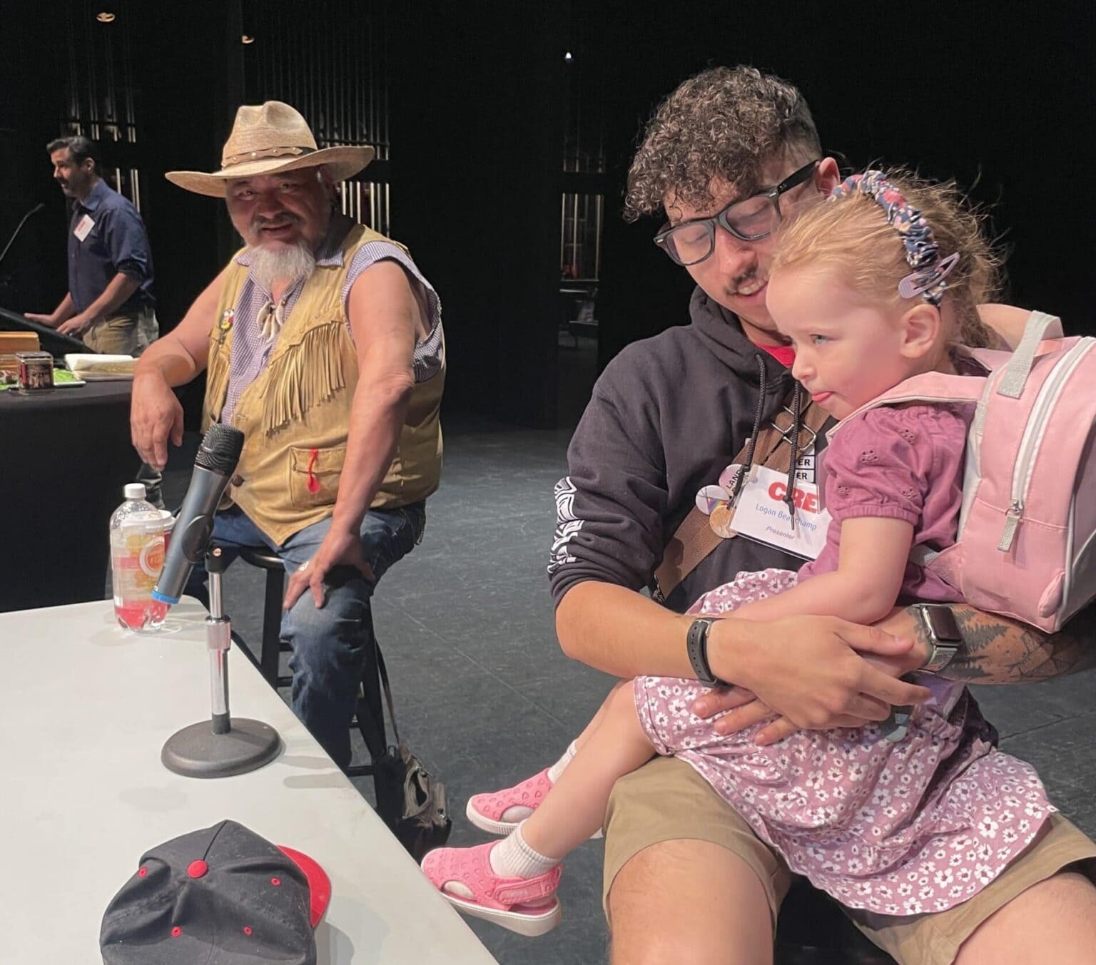 Three people sitting at a table for a panel. One man is wearing a fringe vest and a cowboy hat. The other man wears glasses and a young girl sits on his lap