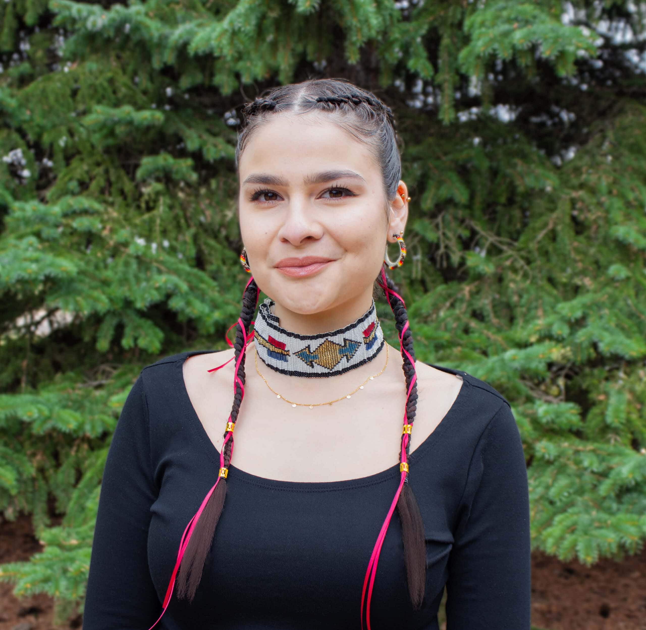 Portrait of Rayna Martens against a background of pine trees. Her hair is braided with red ribbons and she is wearing a beaded choker.