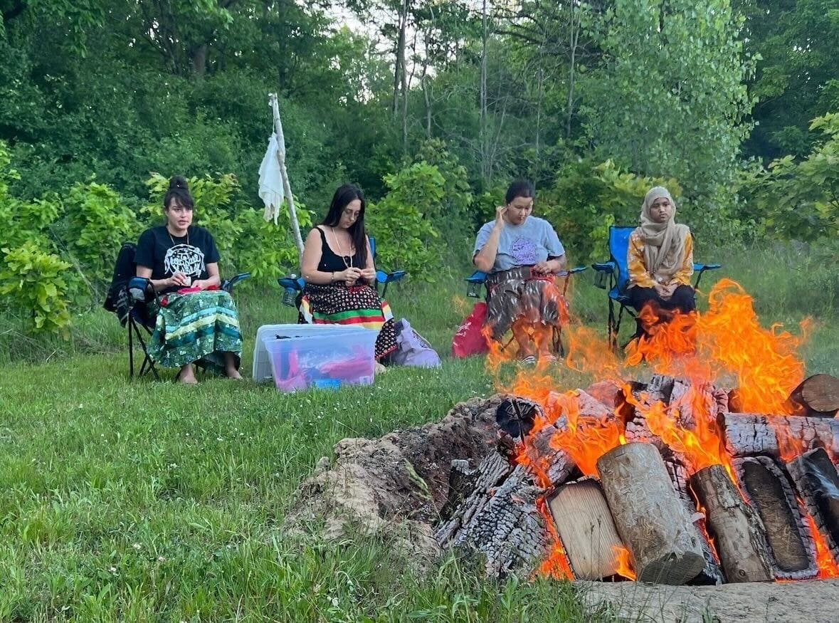 Four youth sitting around a camp fire. They are surrounded by trees and two of them are wearing ribbon skirts.
